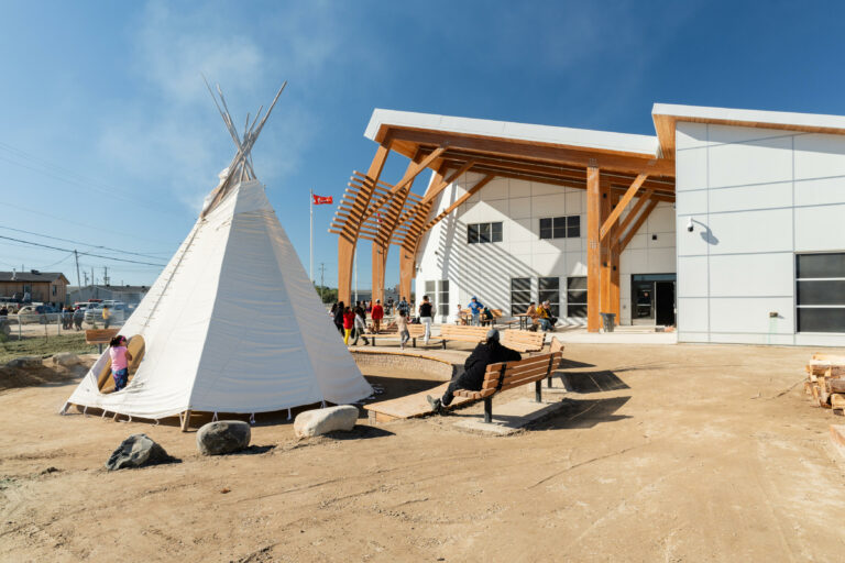 Teepee is set up in front of the Attawapiskat Youth Centre's patio. There are benches around the teepee and on the patio in the background. Community youth are walking around.