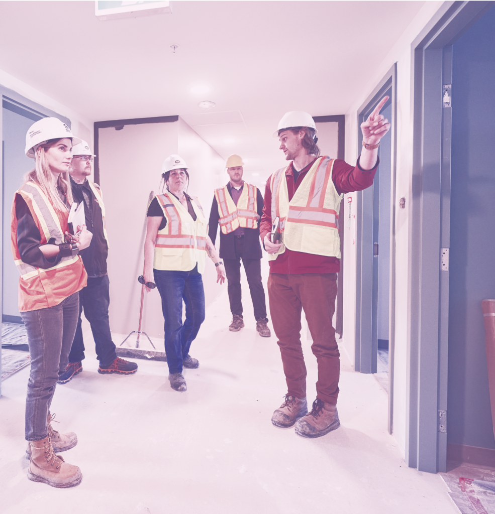 Group of construction workers and architects all wearing safety gear standing in hallway.