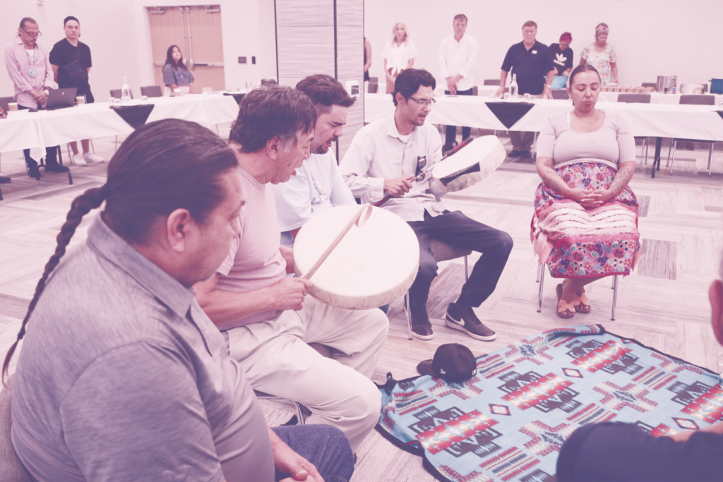 Group of Indigenous people playing drums around a ceremonial blanket.