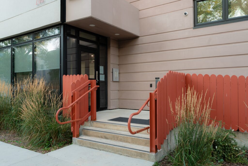 Building's front door with picket fence and wild grass on either side of steps leading to entrance.