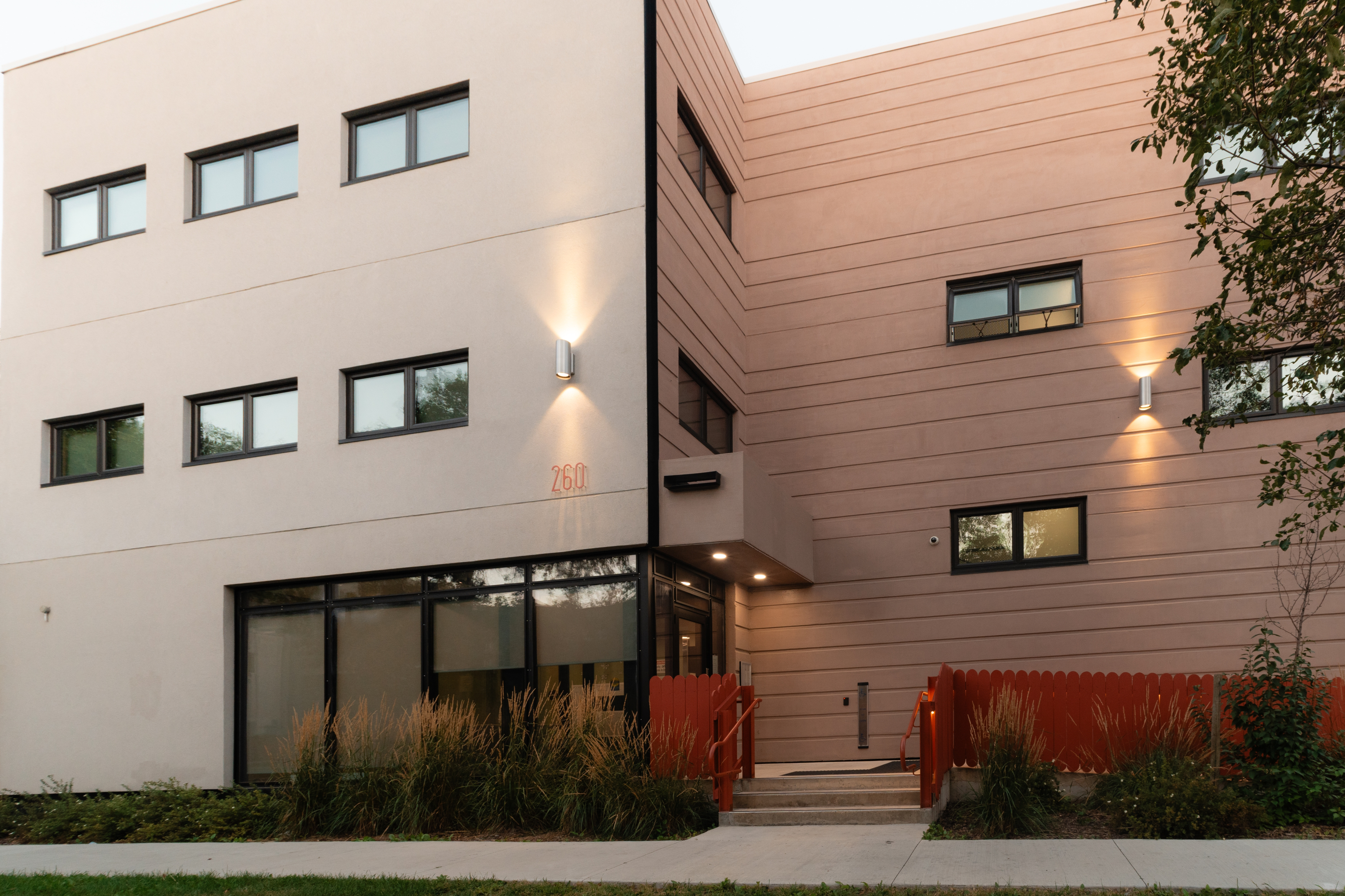 Exterior image of 3-storey apartment building and entry. Burnt orange picket fence in front as well as tall wild grass for landscaping.